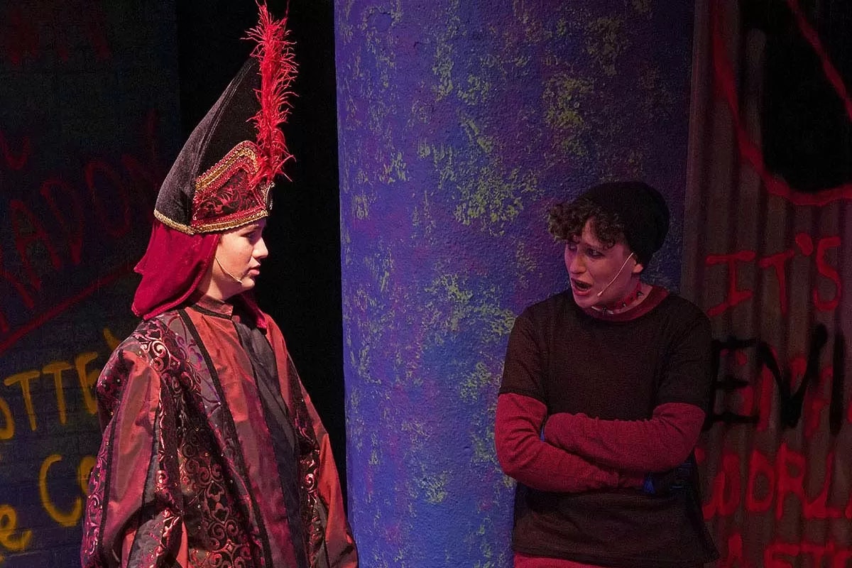Two actors perform on stage; one wears a detailed costume with a tall hat, the other dressed casually, standing near a painted backdrop.