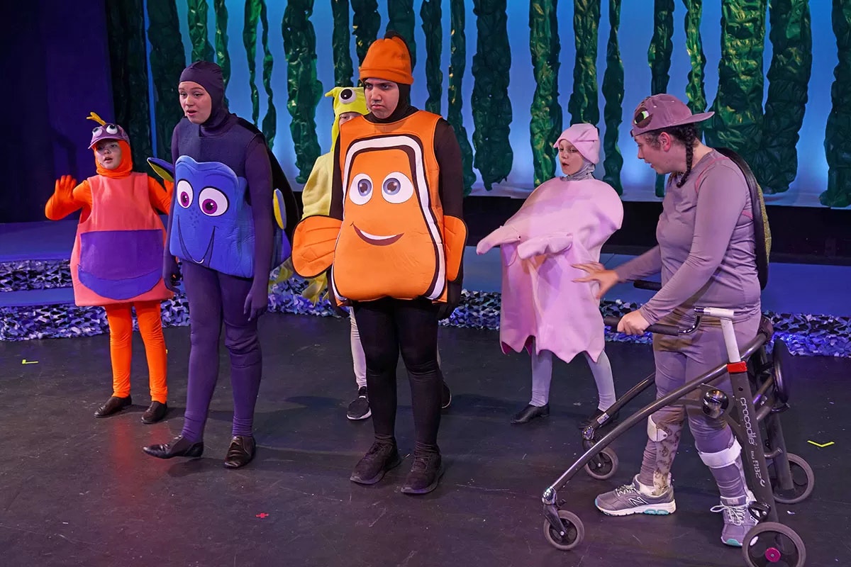 People in colorful fish costumes perform on stage, with seaweed in the background.