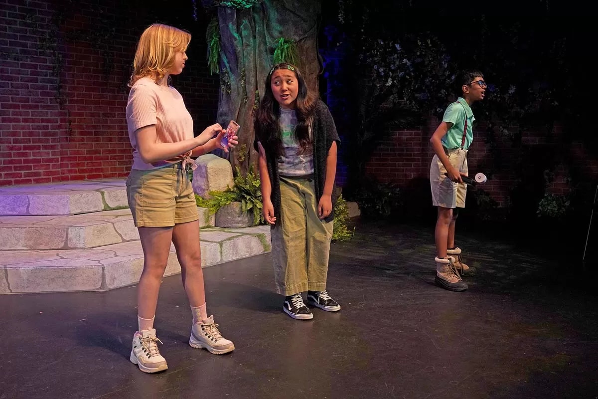 Three people on stage in a forest-themed setting, engaged in a performance.