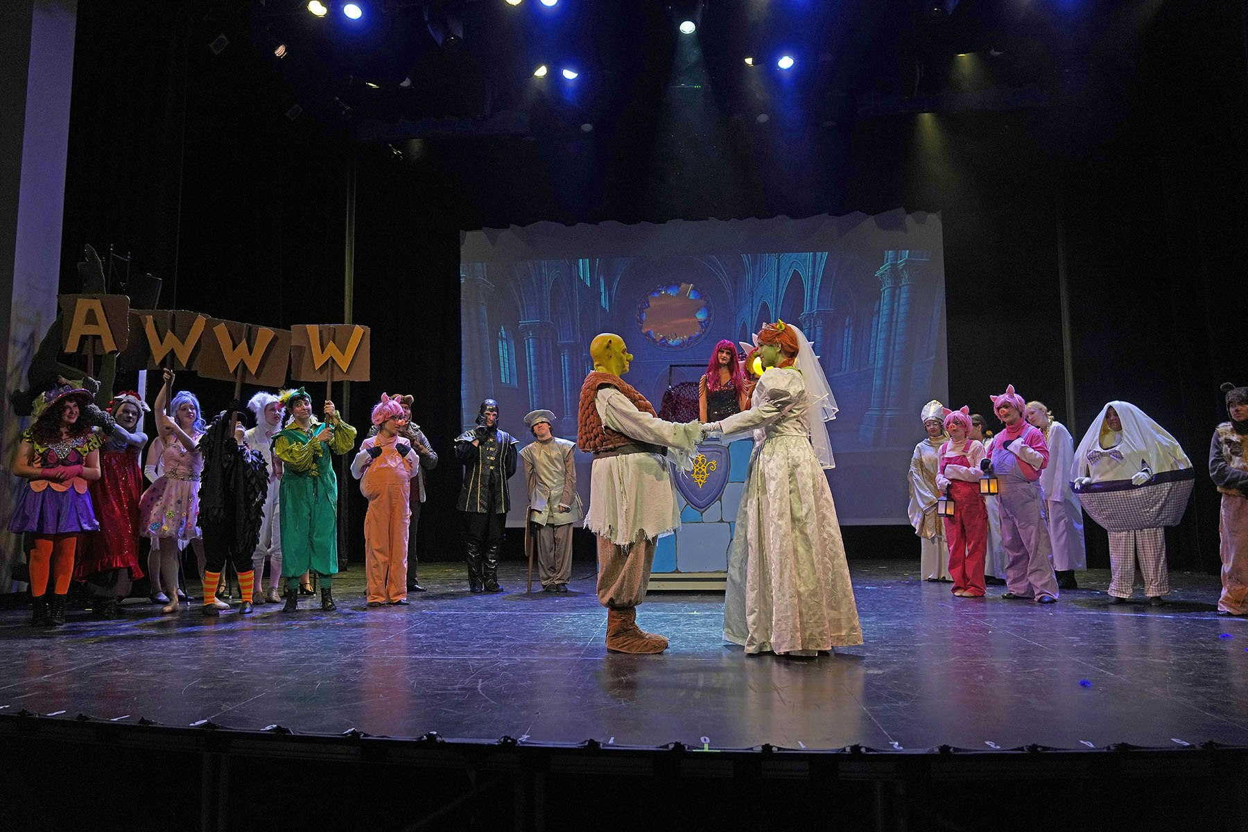 Stage play scene with actors in costumes, including an ogre and a bride, with a backdrop and audience holding "AWWW" signs.
