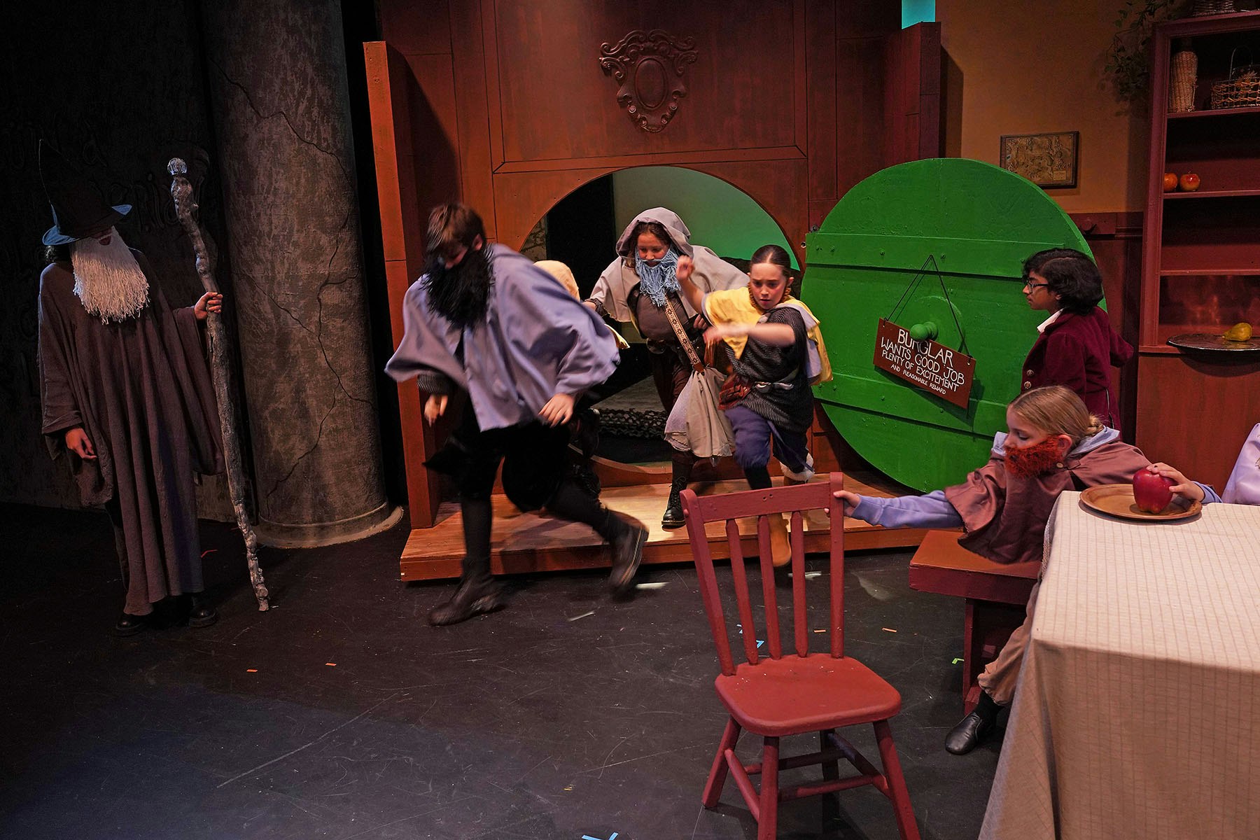 People in costumes on a stage, depicting a scene with a large door, wizard, and medieval characters.
