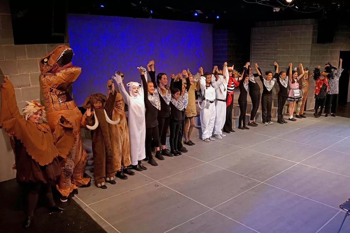 A group of performers in costumes, including animals, on stage holding hands, likely after a show.