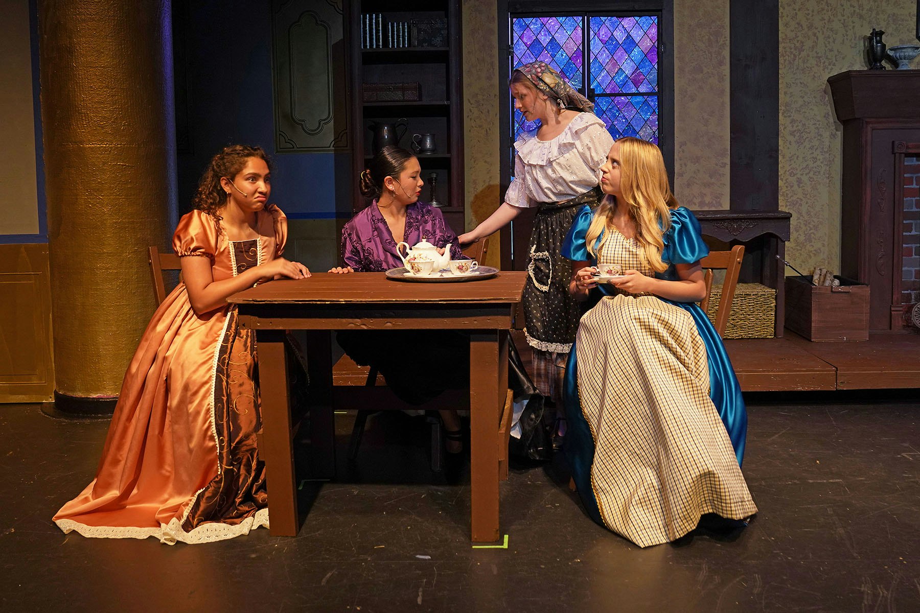 A scene from a play features four women in period costumes around a table, engaged in conversation with tea set.
