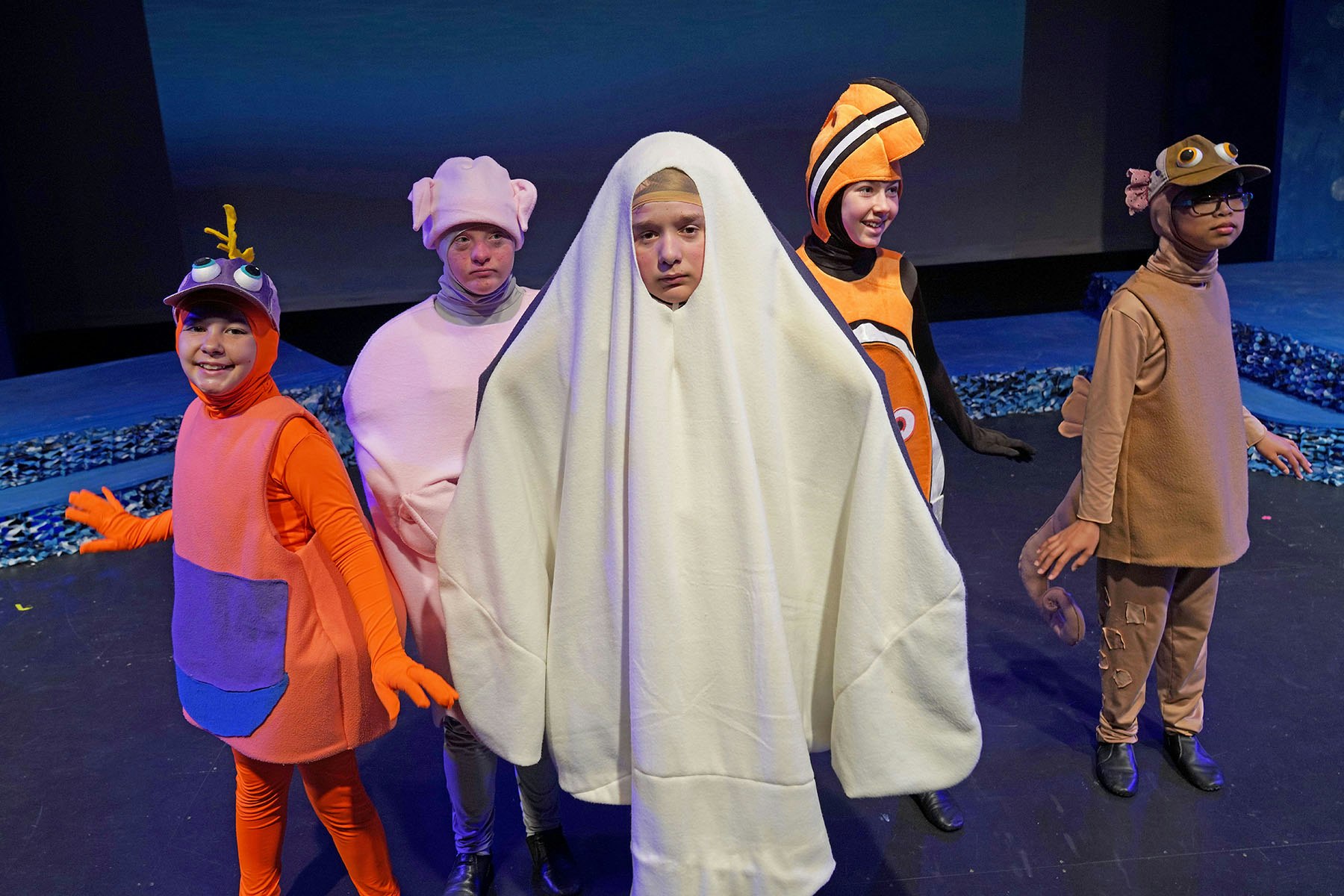 Five kids in colorful animal and sea creature costumes on stage, one dressed as a ghost.