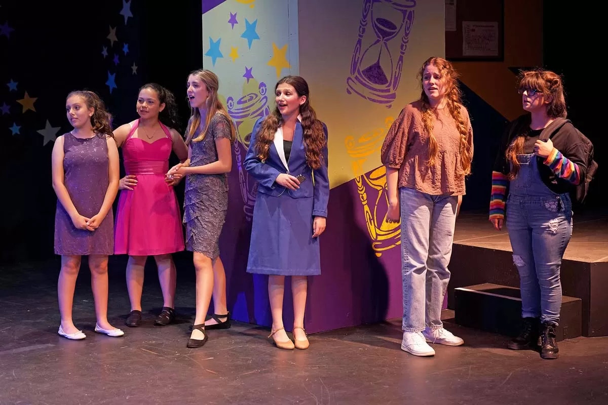 A group of six people are standing on a stage, dressed in various outfits, with colorful star and hourglass decorations in the background.