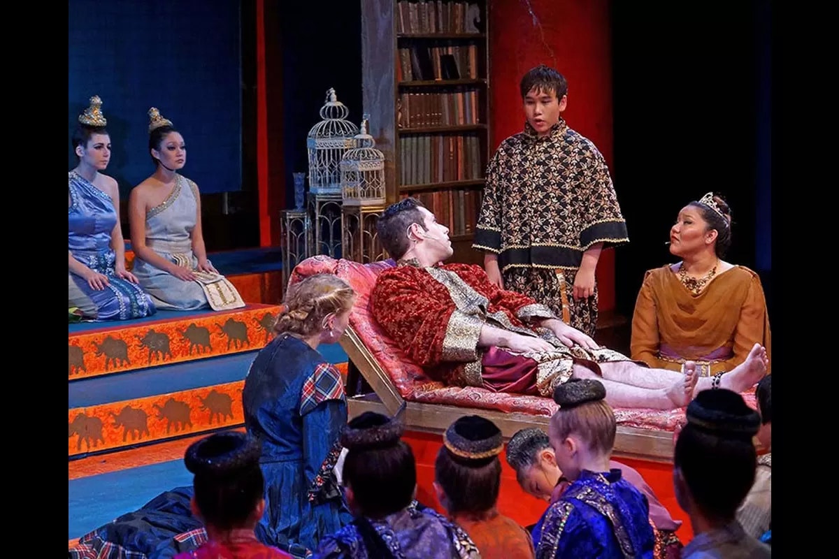 A theatrical scene with actors in elaborate costumes, a person lying on a couch, and others surrounding them, engaged in a dialogue.