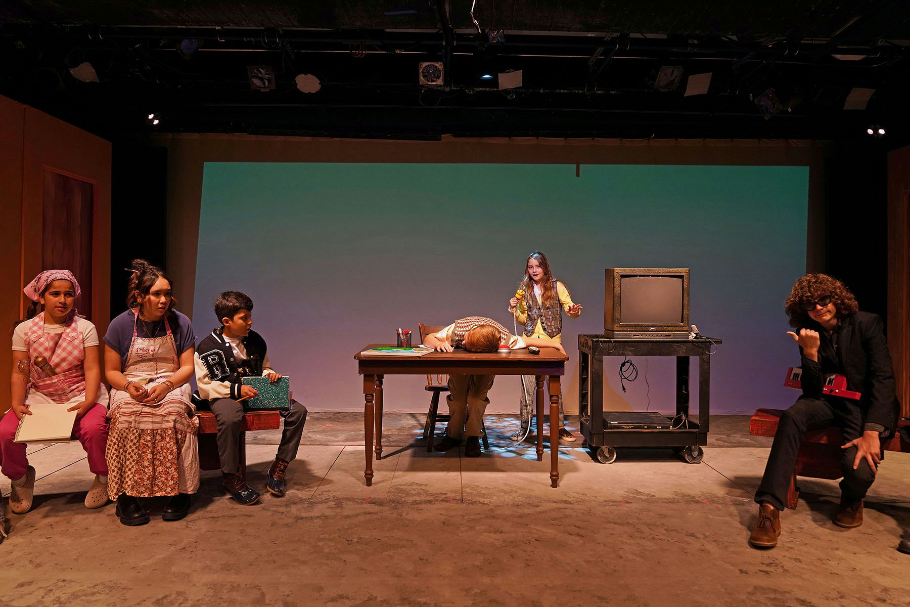 A theatrical scene featuring six performers: four seated, one at a desk acting, and a vintage TV on display.