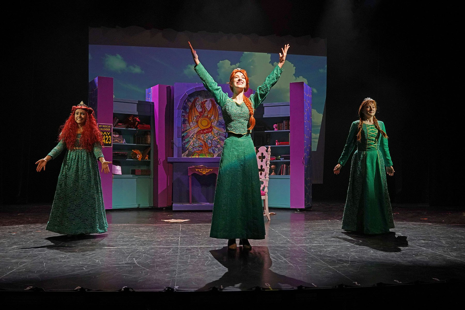Three performers in green costumes on stage, with colorful shelves and a stained glass backdrop.