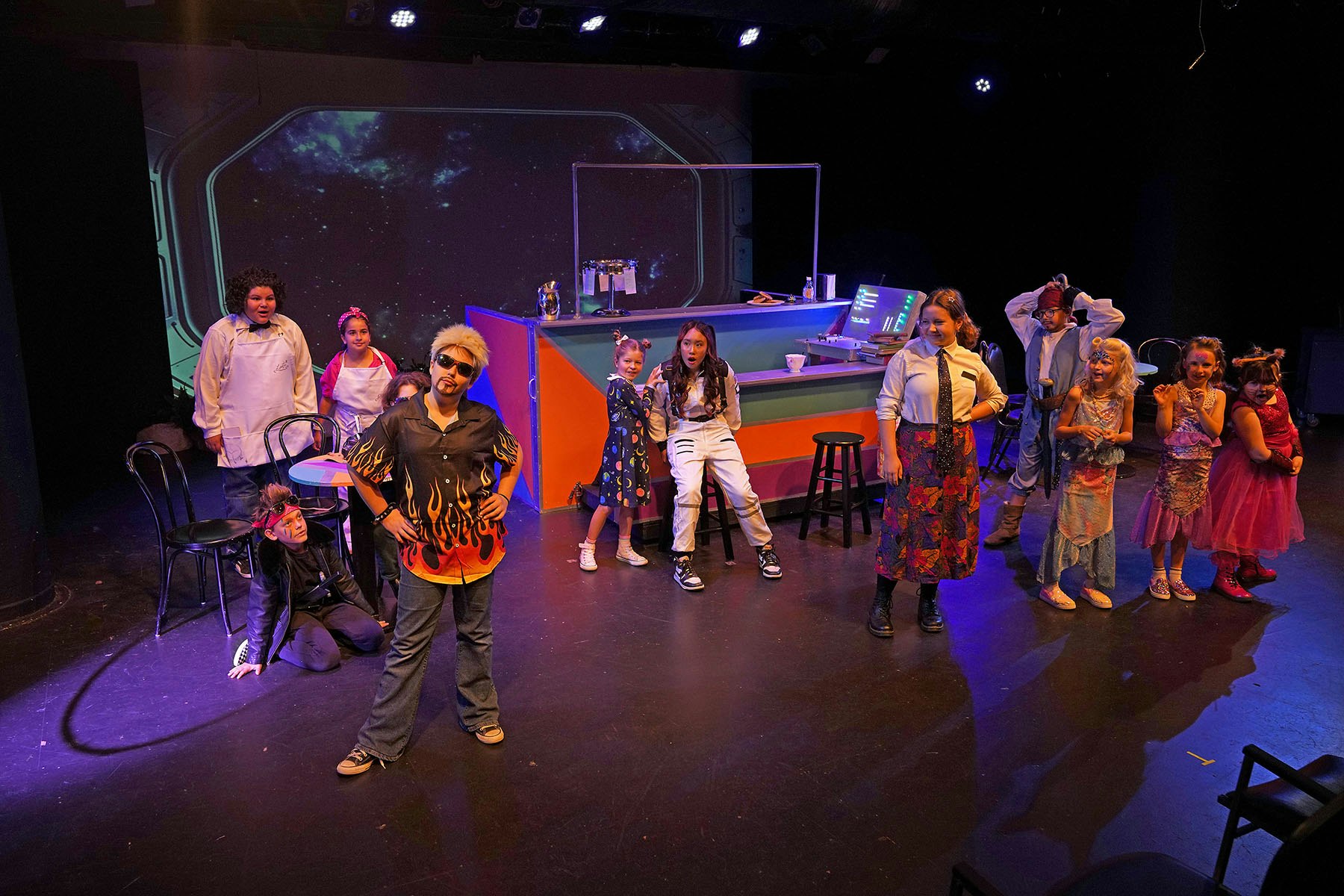A group of people in colorful costumes on a stage set resembling a space-themed diner.
