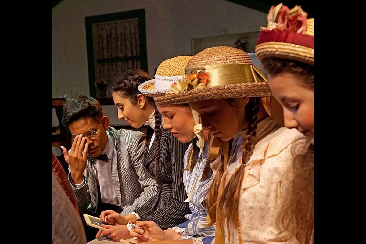 A group of people in vintage attire, some wearing straw hats, sitting closely and focused on an unseen point.