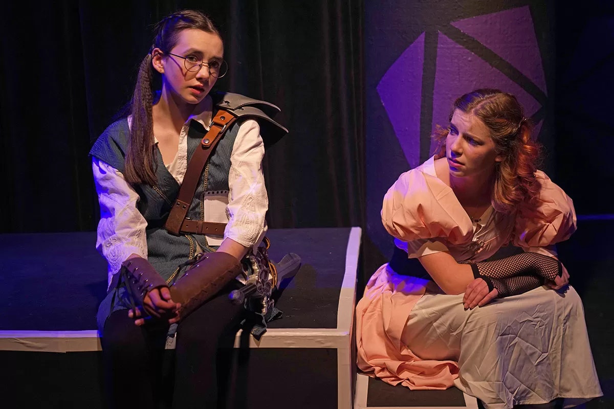 Two people in costumes, sitting on stage, with serious expressions.