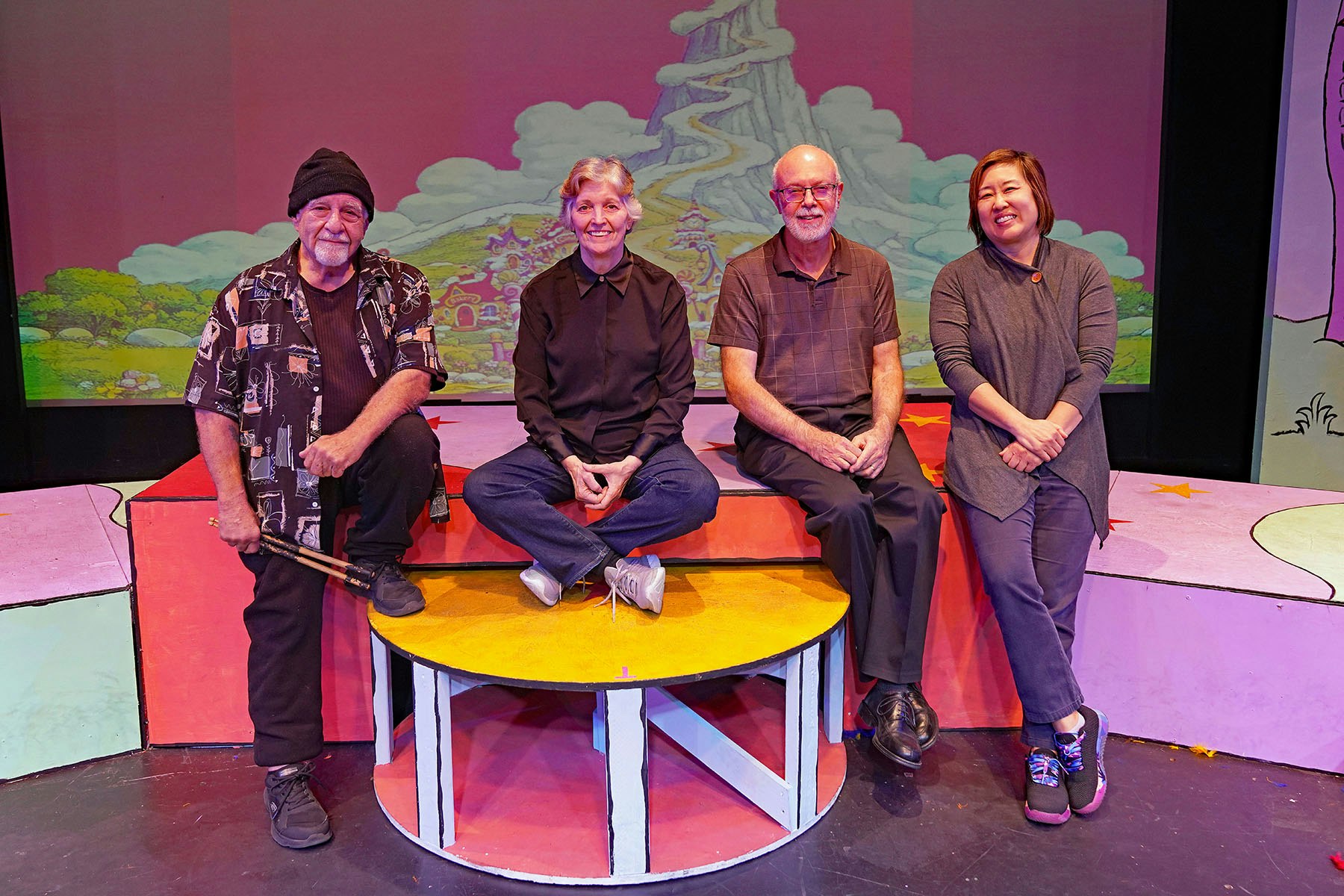 A group of four individuals poses together on a colorful stage set, with a whimsical background depicting a cartoon landscape.
