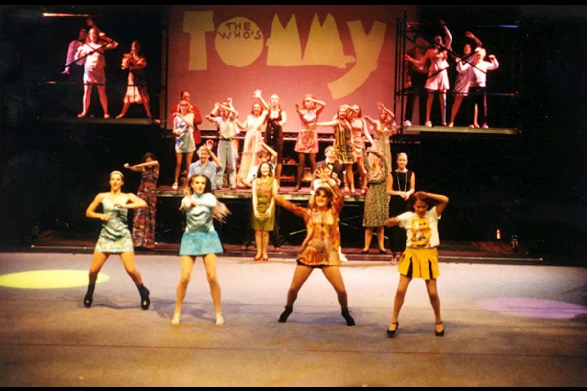 A stage performance with dancers and a backdrop reading "The Who's TOMMY." Several performers are in various colorful costumes.