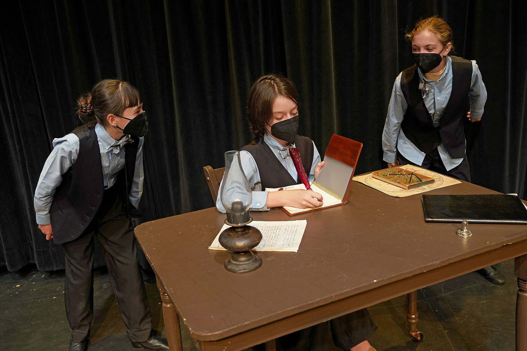 Three people in suits and masks, one writing at a table with documents and an old lamp, others observing.