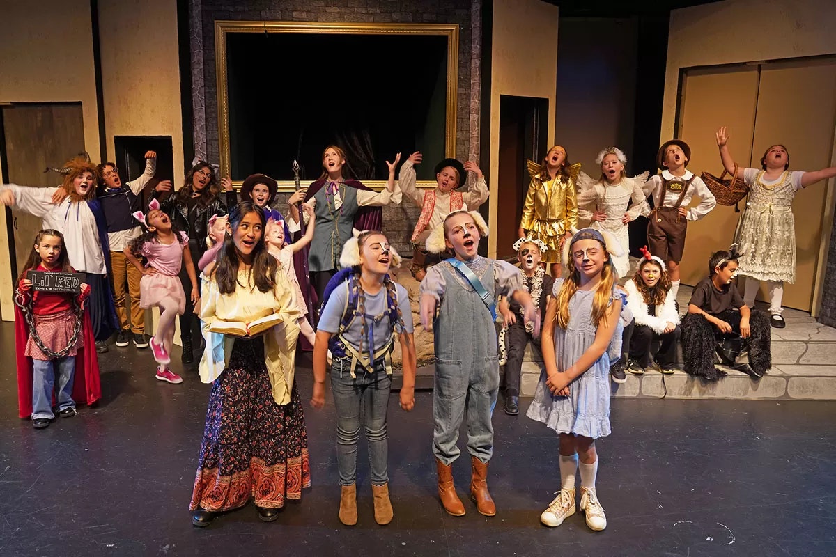 A group of children in colorful costumes performing on stage, some holding props, possibly in a play or musical.