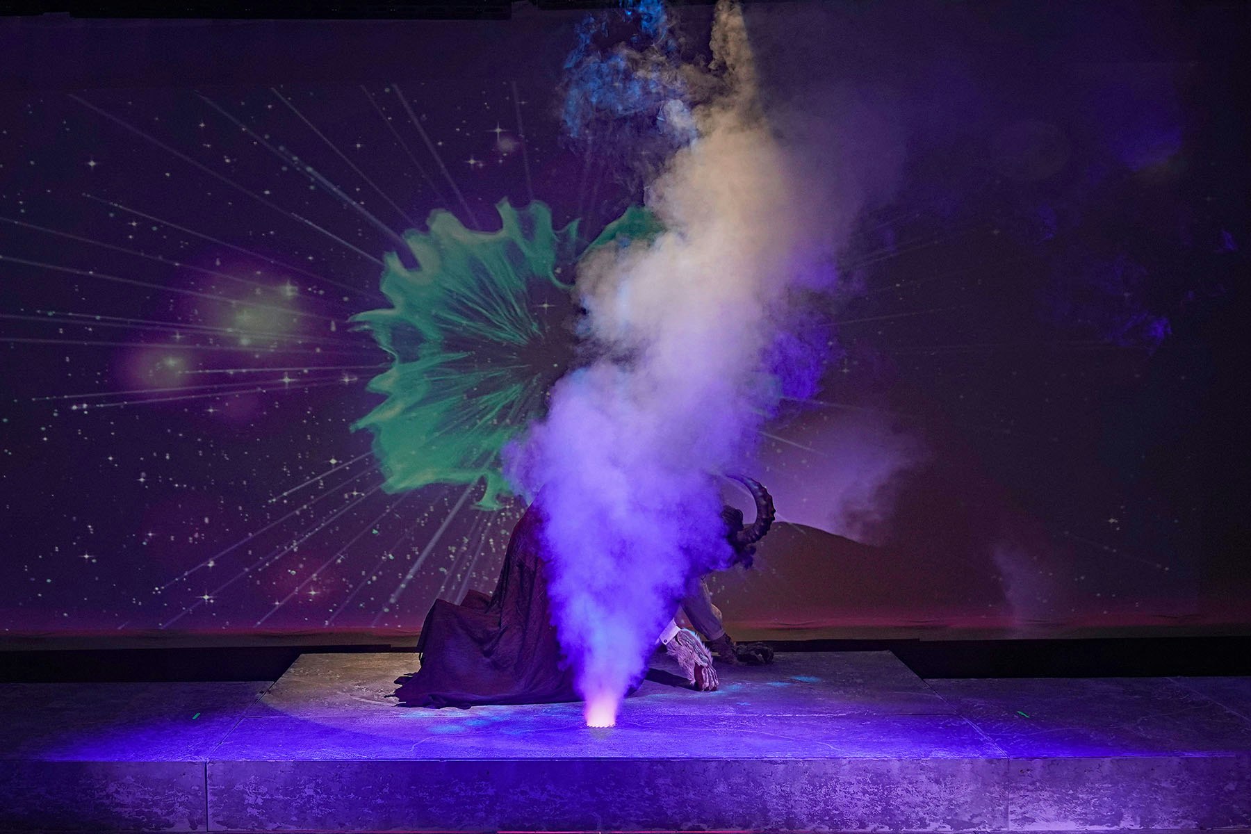 A dramatic stage scene with smoke, a figure with horns kneeling, and a cosmic backdrop with starry effects.