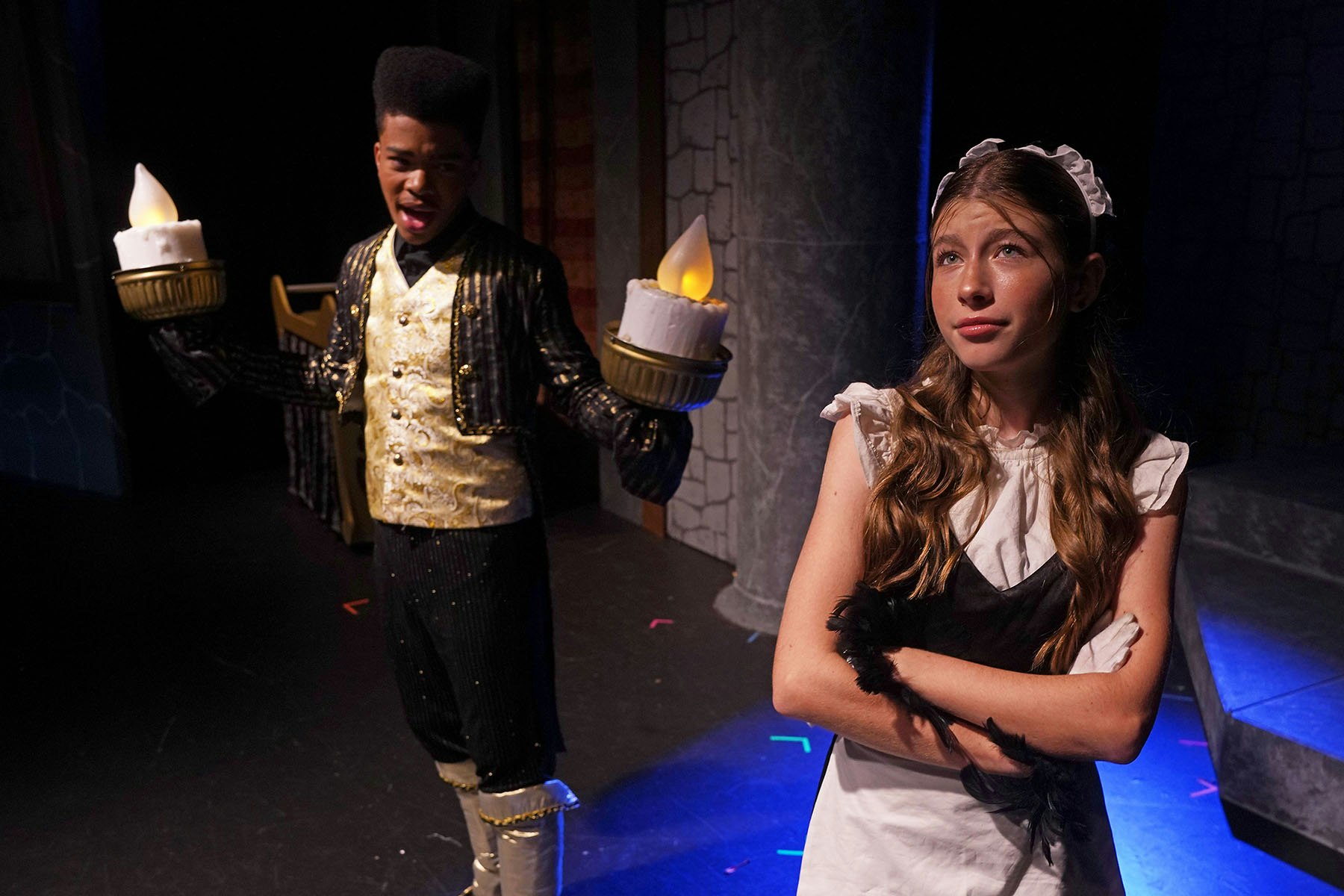 Two people in costume on stage: one as a candlestick singing, the other with crossed arms dressed as a maid.
