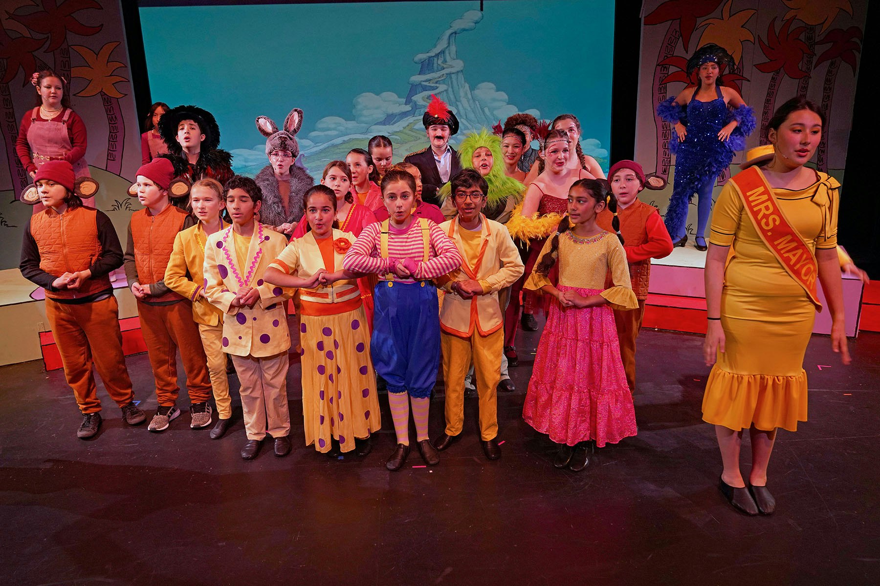 A colorful children's theater performance with a large group of kids in vibrant costumes on stage.