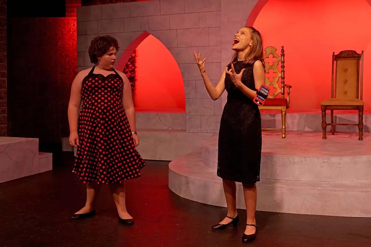 Two women in dresses perform on stage with a dramatic, red-lit backdrop and two chairs.