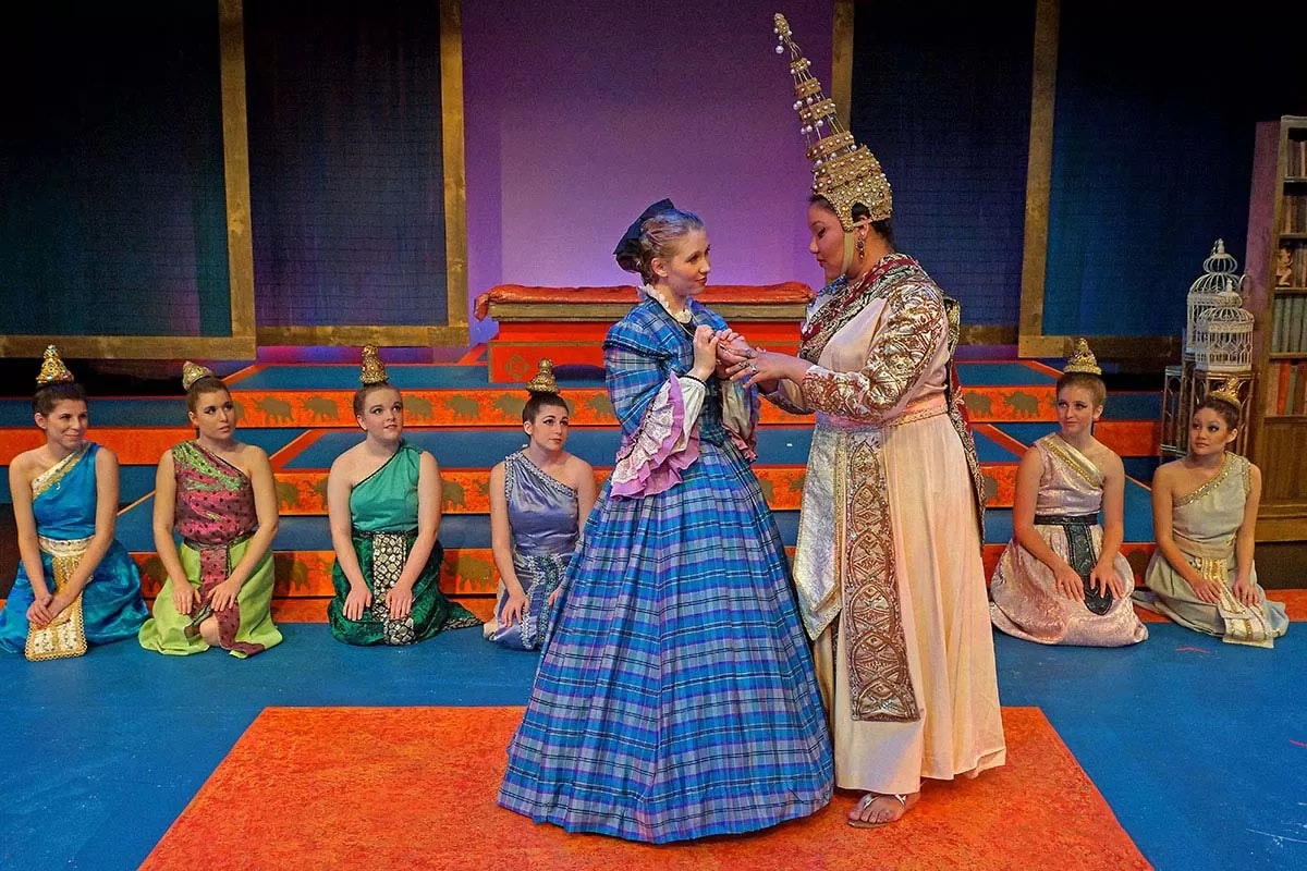 A theatrical scene with people in elaborate costumes, including one in a blue dress and another in ornate traditional attire, others kneeling.
