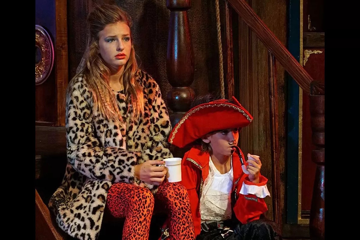 Two people in costume sit; one in a leopard coat with a white cup, the other dressed as a pirate with a pipe.