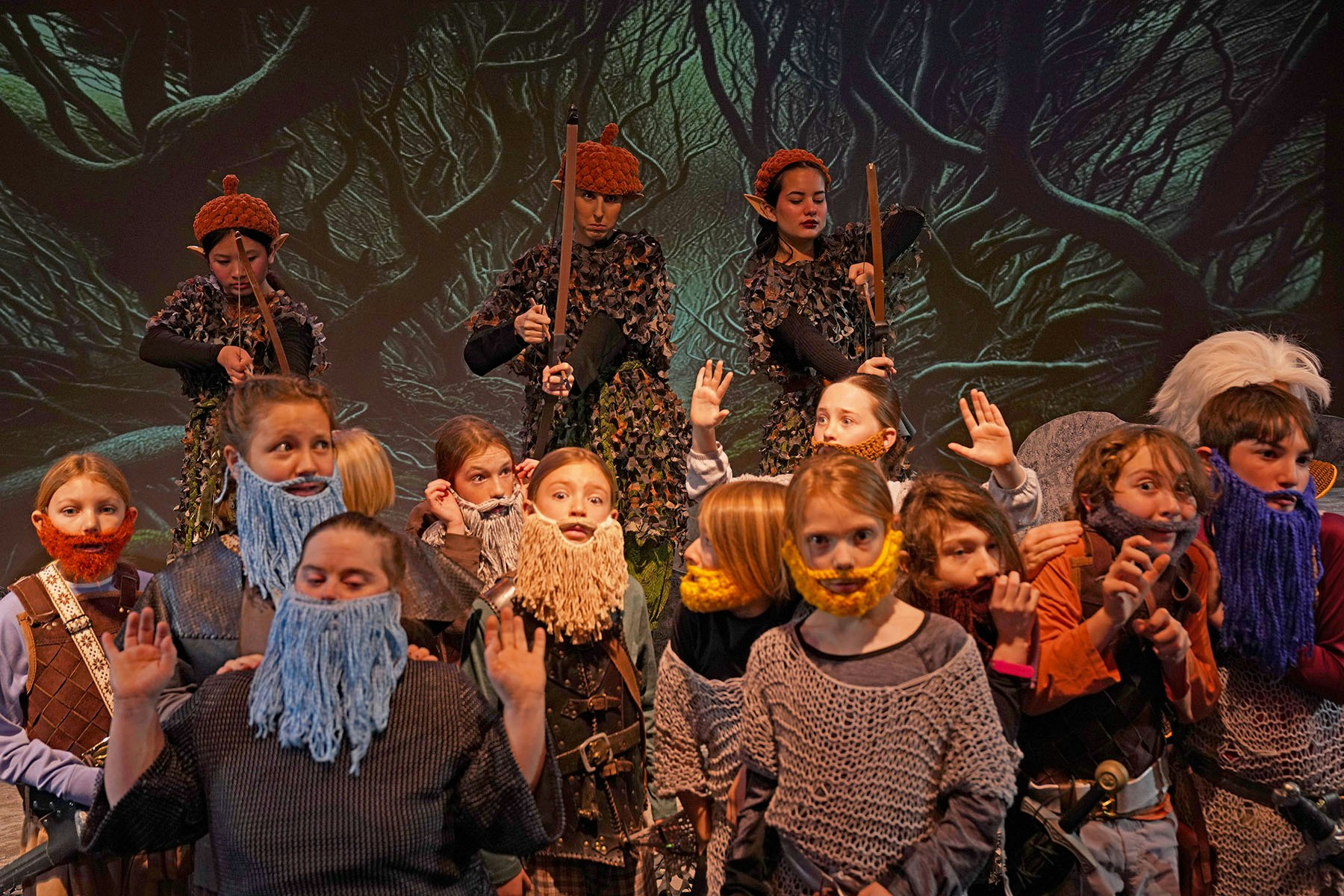 Children in fantasy costumes, some with yarn beards and elf ears, pose on stage with a forest background.