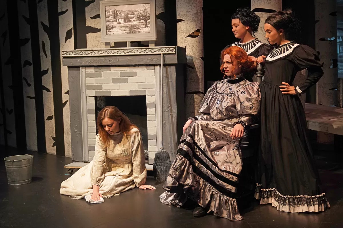 Four women in period costumes; one kneels by a fireplace, others stand around, with a painting above.