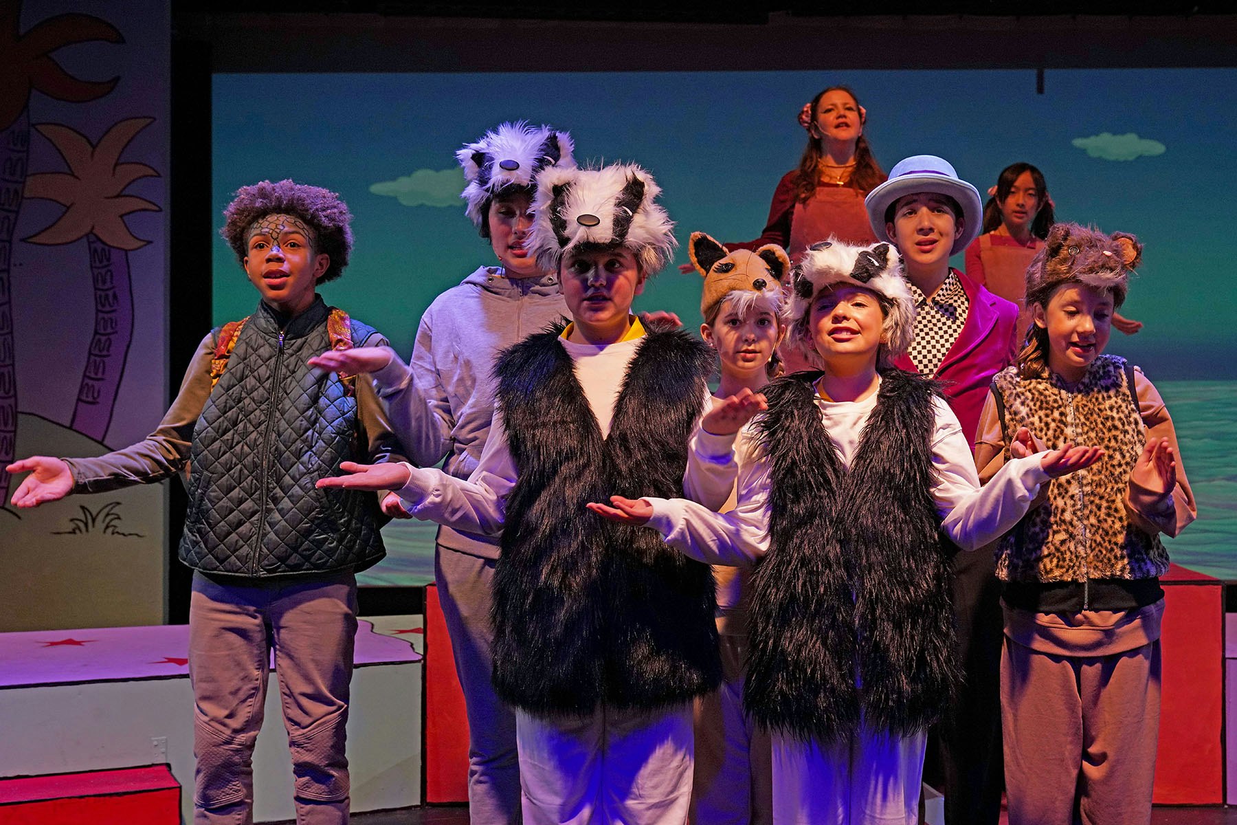 A group of young performers dressed in animal-themed costumes, singing and acting on stage with a colorful backdrop.