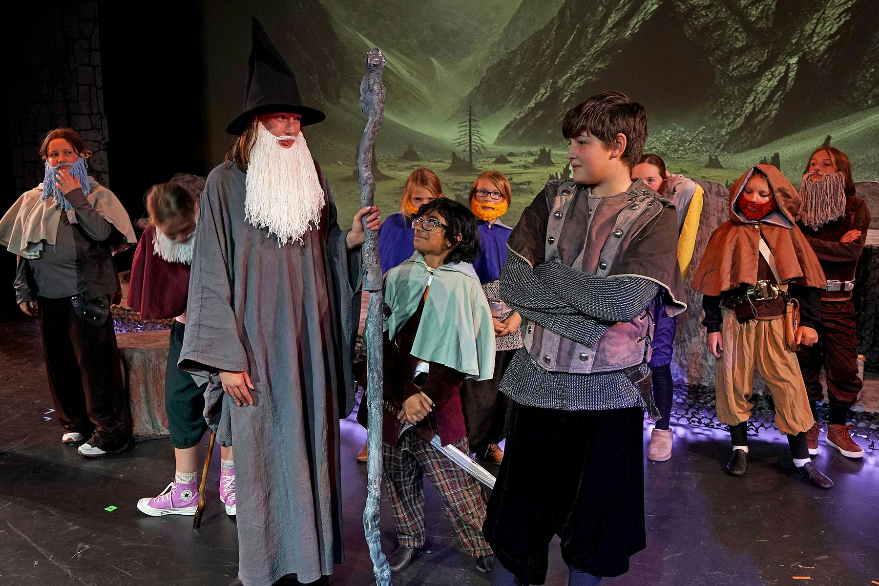 A group of kids dressed in fantasy costumes on stage, resembling characters from an adventure story, with mountains in the background.