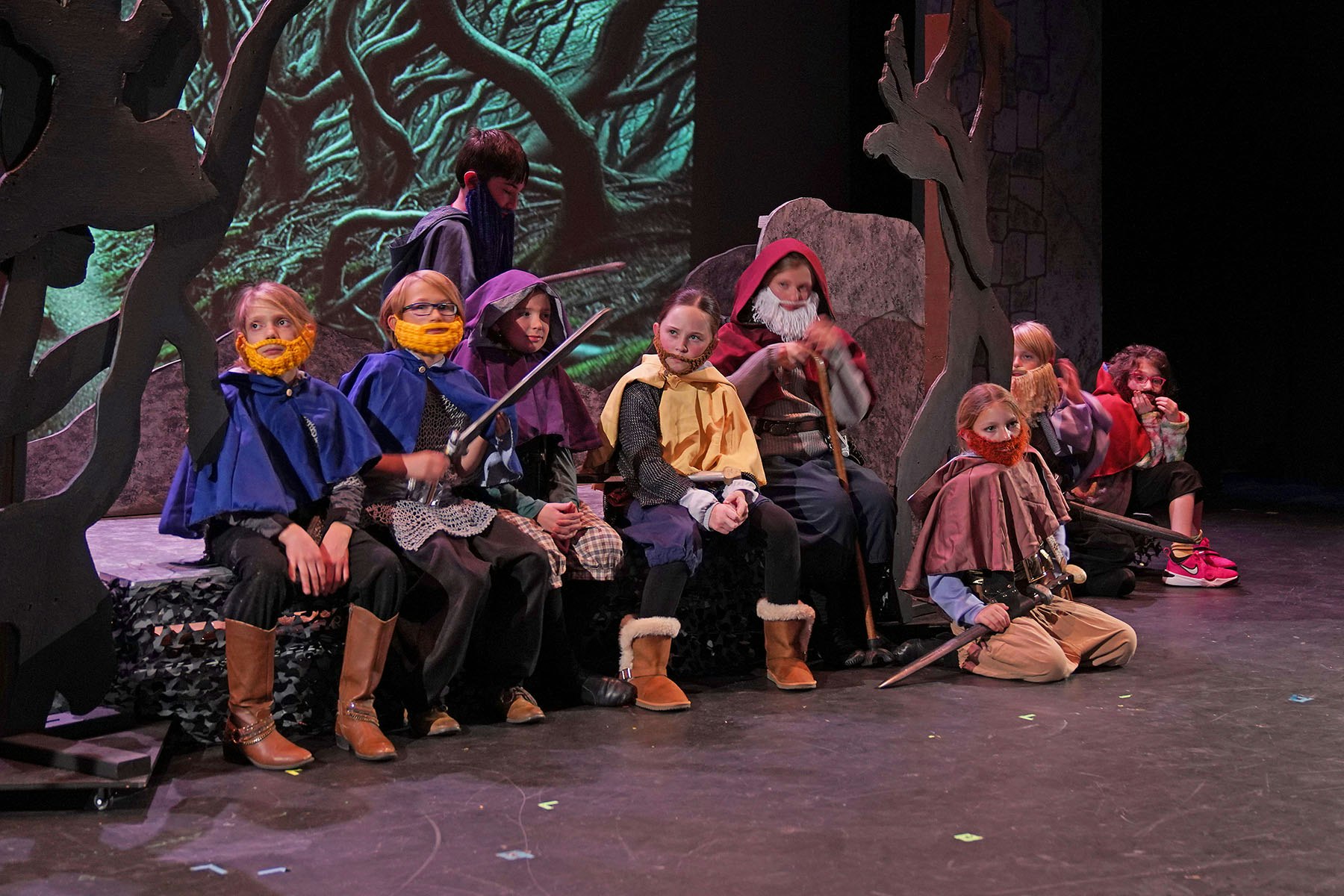 Children in fantasy costumes with beards and capes on a stage set with trees and a mystical backdrop.