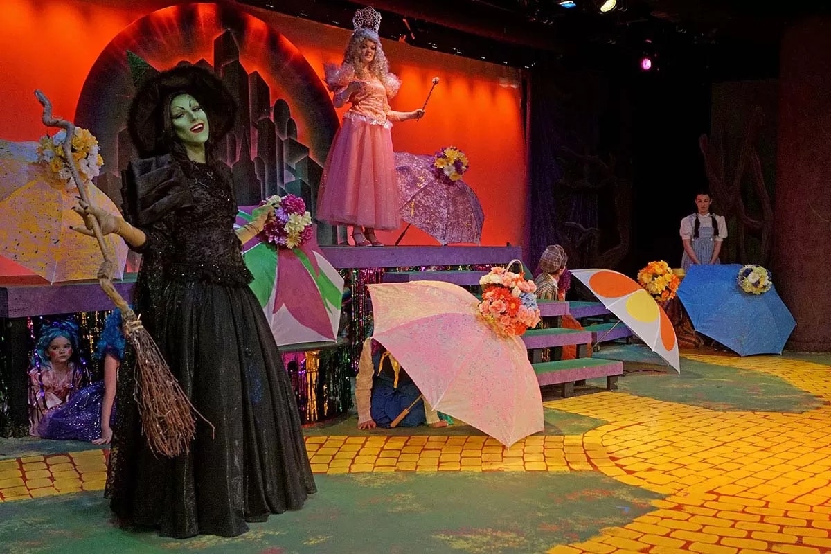 Stage play scene with characters in colorful costumes, including a green witch and a pink fairy, with umbrellas and a painted backdrop.