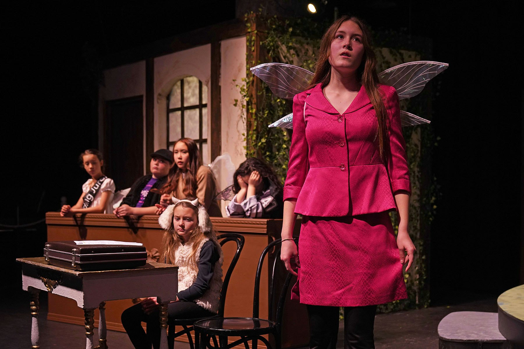 A young woman in a pink outfit with wings stands on stage, surrounded by seated people, one wearing animal ears, in a theatrical setting.