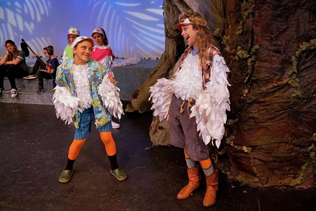 Children in bird costumes performing on stage, surrounded by a whimsical forest set.