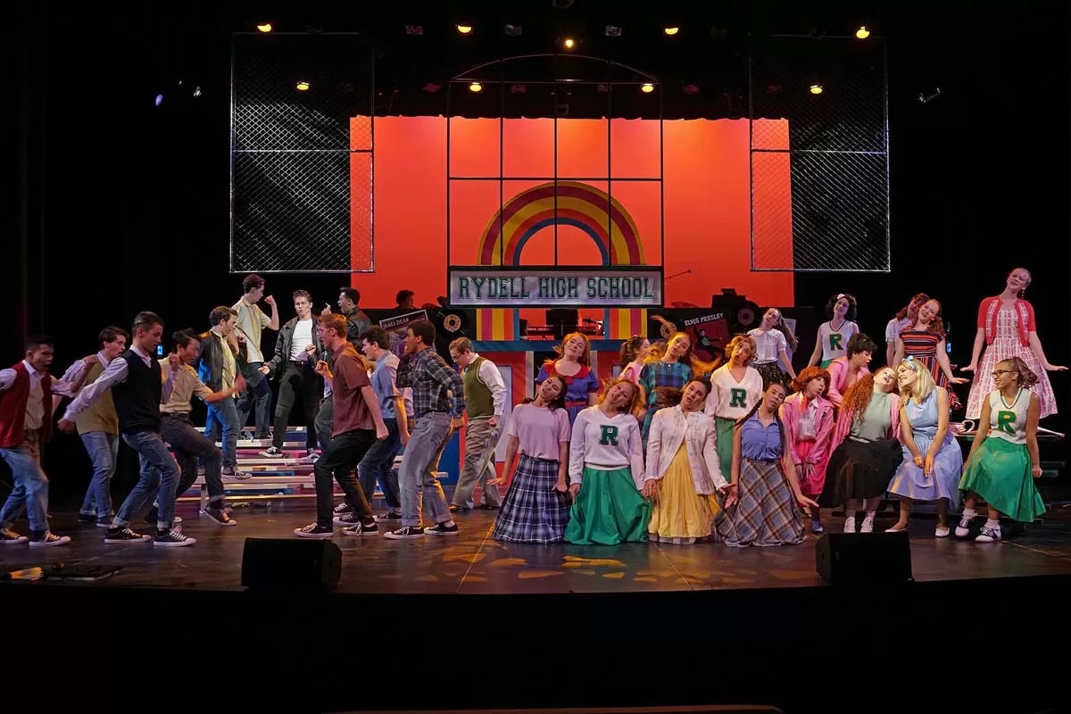 A theatrical performance with a group of people on stage, featuring "Rydell High School" and colorful 1950s-style costumes.