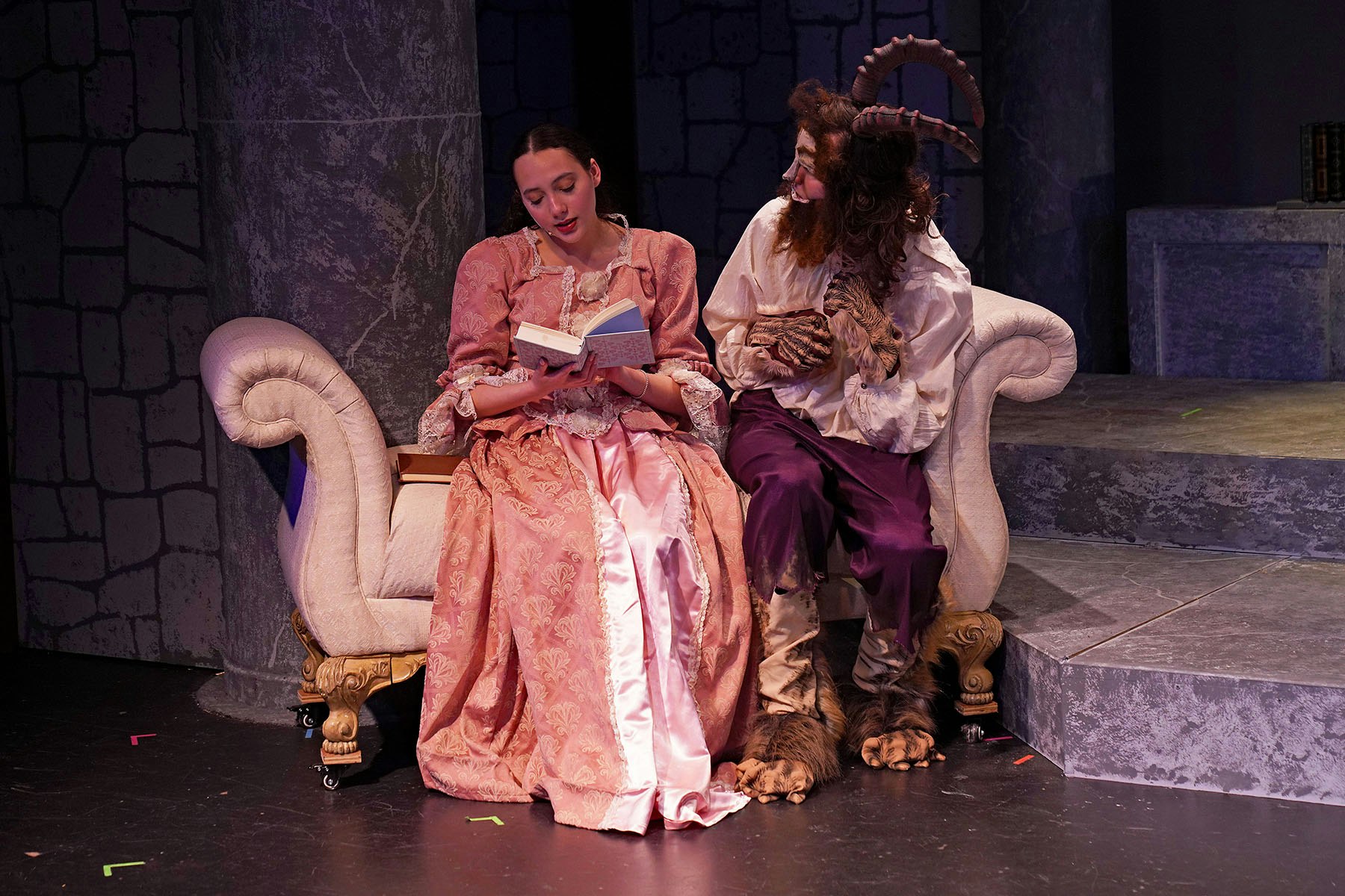 A man in a beast-like costume and a woman in a pink dress sit on a couch; she is reading a book.