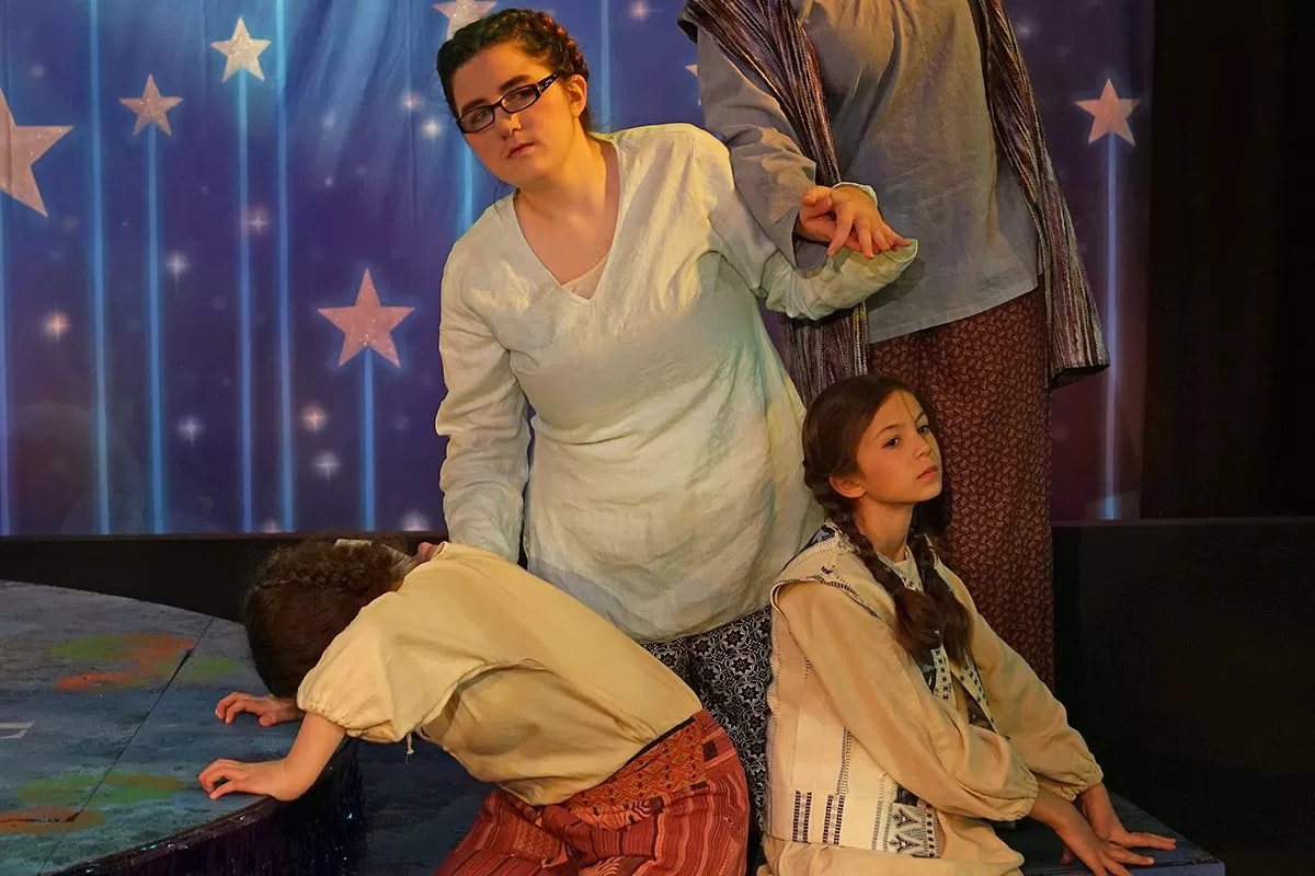 Three people in a play setting. One person appears thoughtful, one is bent over, and another sits with a serious expression. Stars backdrop.