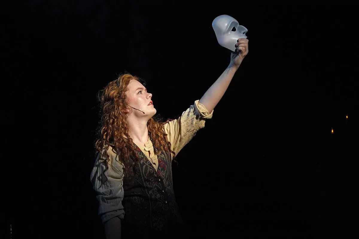 A person on stage holds up a white mask, looking upwards with dramatic lighting.