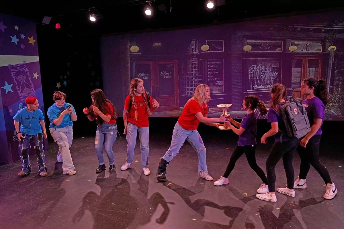 A group of people in colorful shirts acting on stage, one hands an object to another.