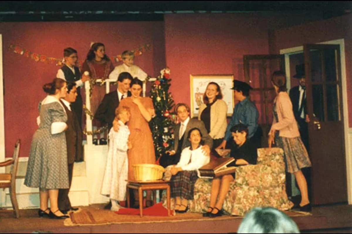 A group of people in vintage attire on stage, possibly in a play, gathered near a decorated Christmas tree in a living room setting.