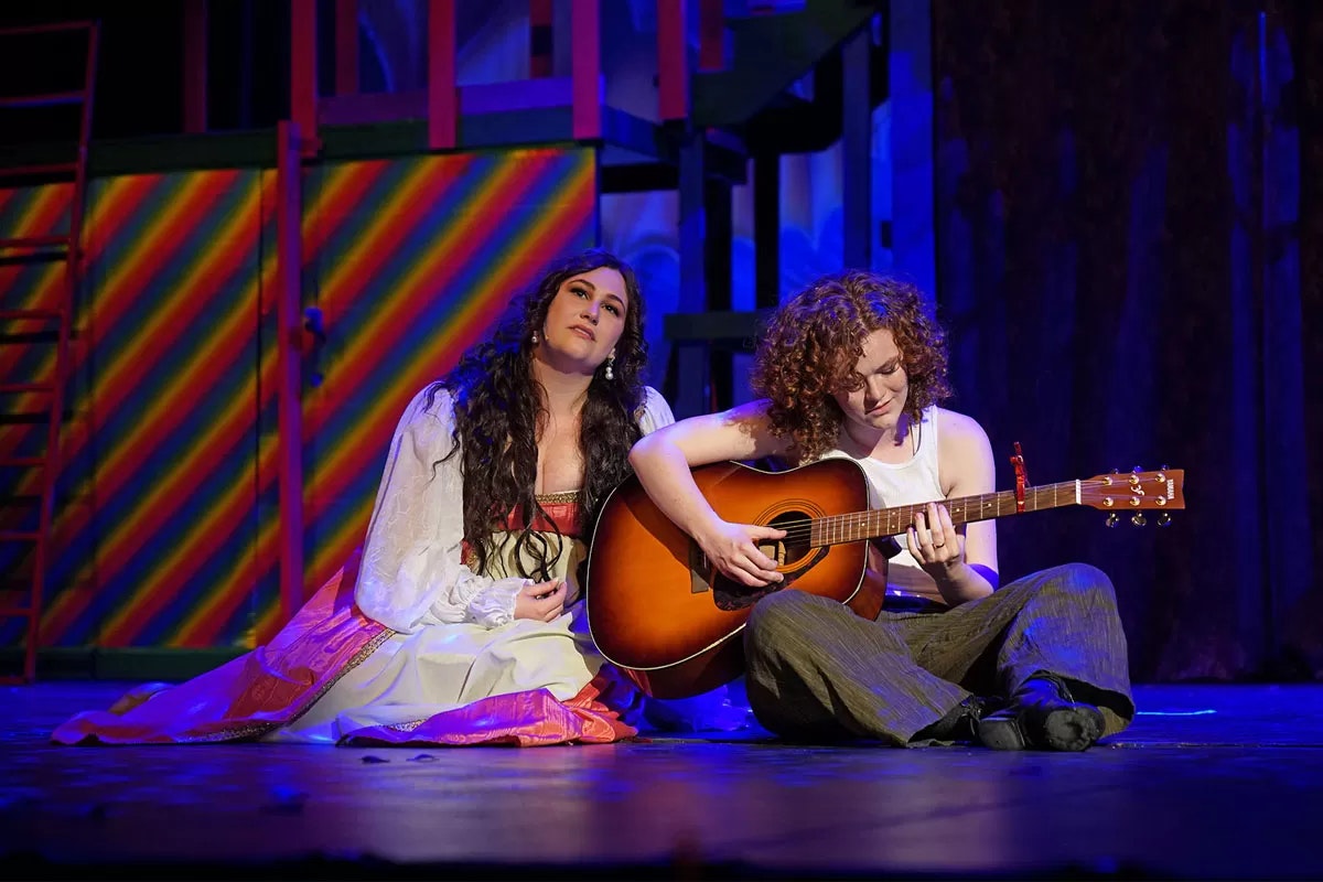 Two people sit on a stage; one plays a guitar, the other listens. The background has rainbow stripes.