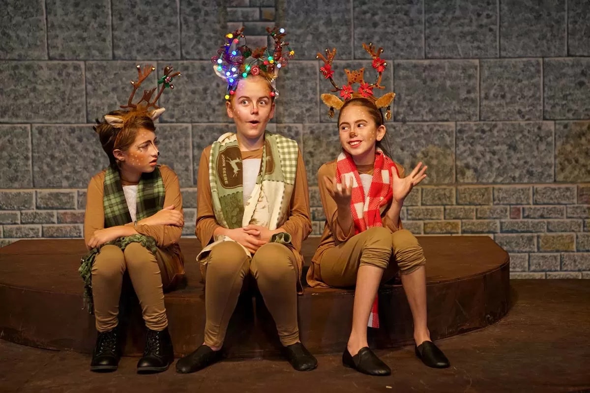 Three children dressed as reindeer with antlers and scarves sit on a stage, showing playful and expressive poses.