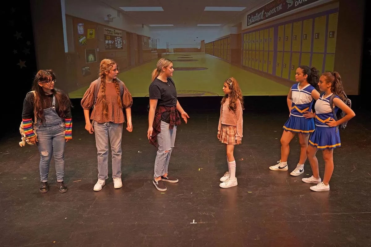 A group of people are performing on stage with a school hallway projected behind them.