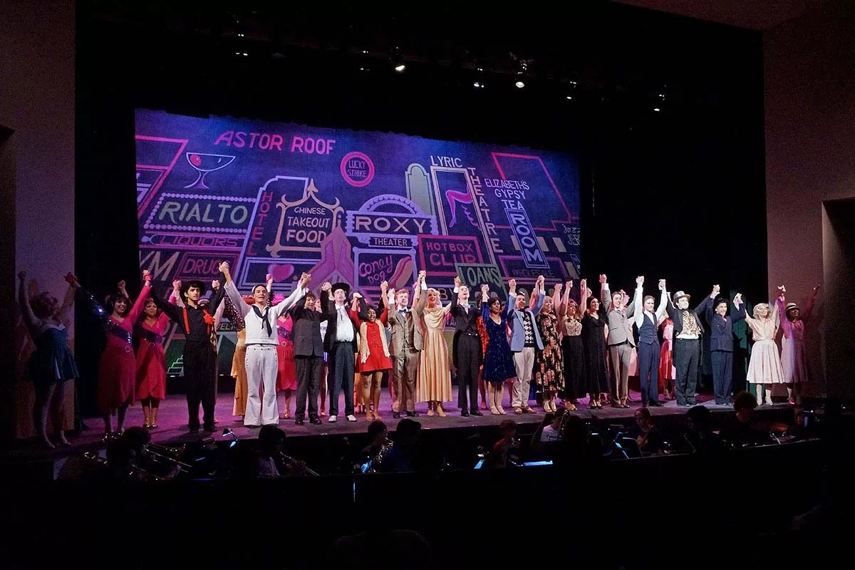 A group of performers in costume takes a bow on stage against a colorful backdrop with theater-themed graphics.