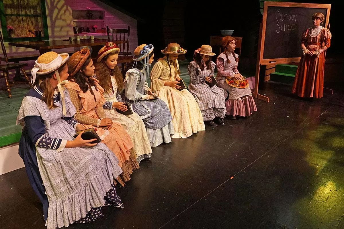 A group of girls in vintage dresses and hats are seated, holding books, with "Sunday School" written on a blackboard.
