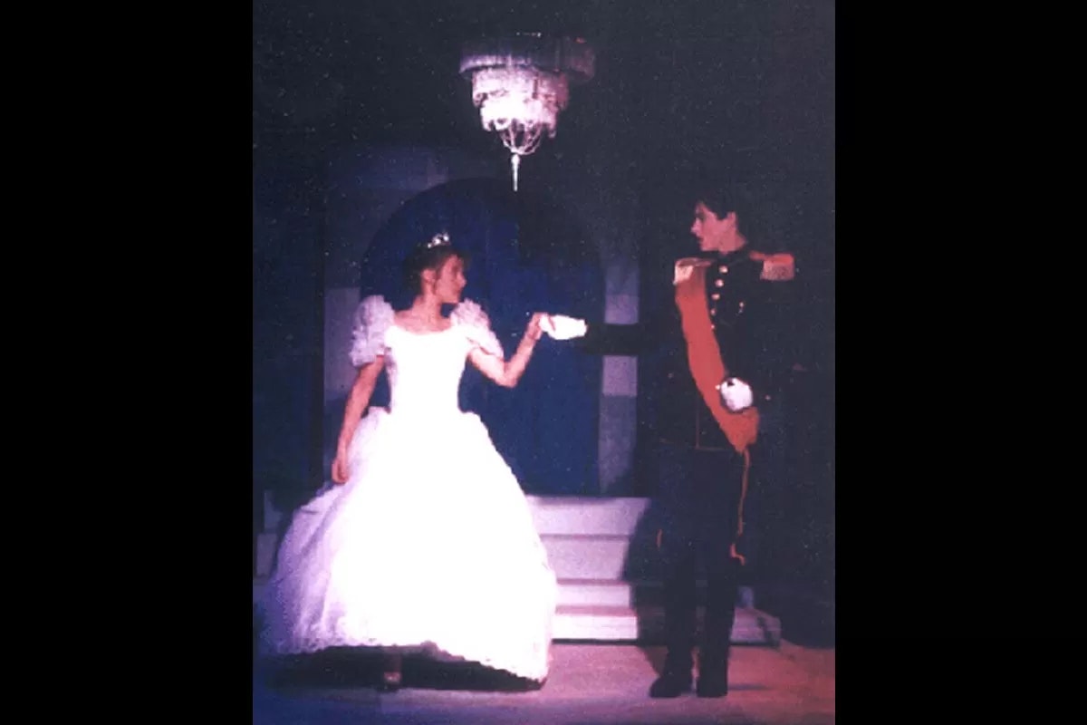 A woman in a white gown and a man in a formal uniform are standing under a chandelier, possibly in a dance pose.