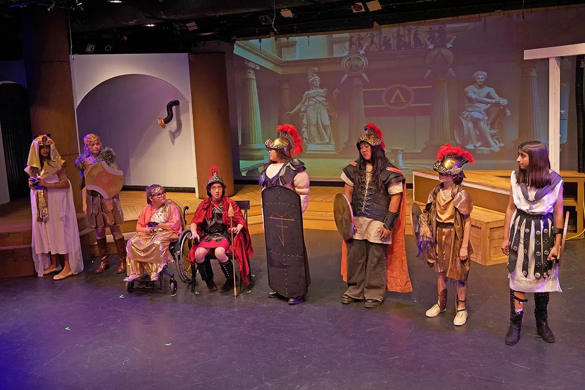 People in historical-themed costumes on stage, with a backdrop of classical architecture.