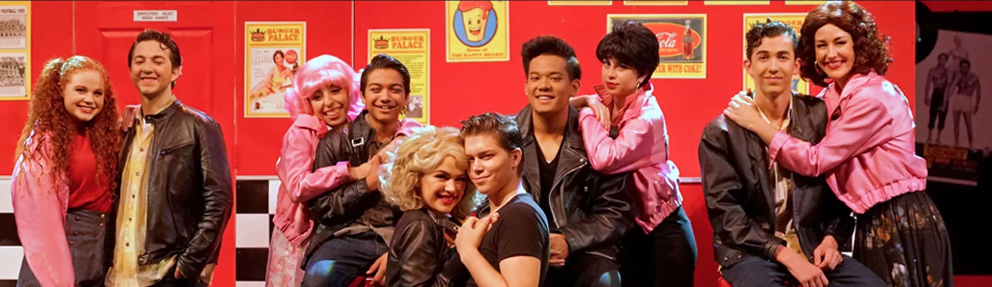 A group of people in costumes pose together in front of a red backdrop with "Burger Palace" signs.