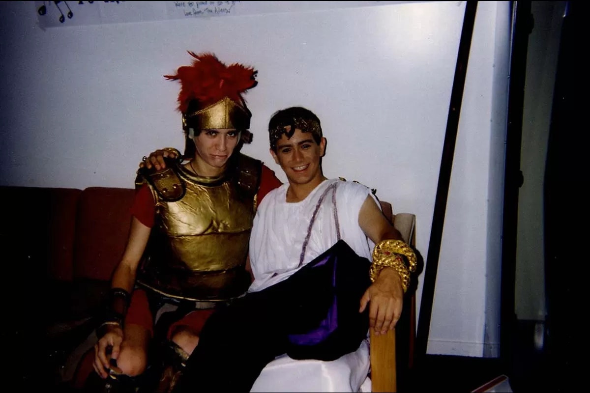 Two people are in Roman-themed costumes, one in armor and the other in a toga, sitting together indoors.