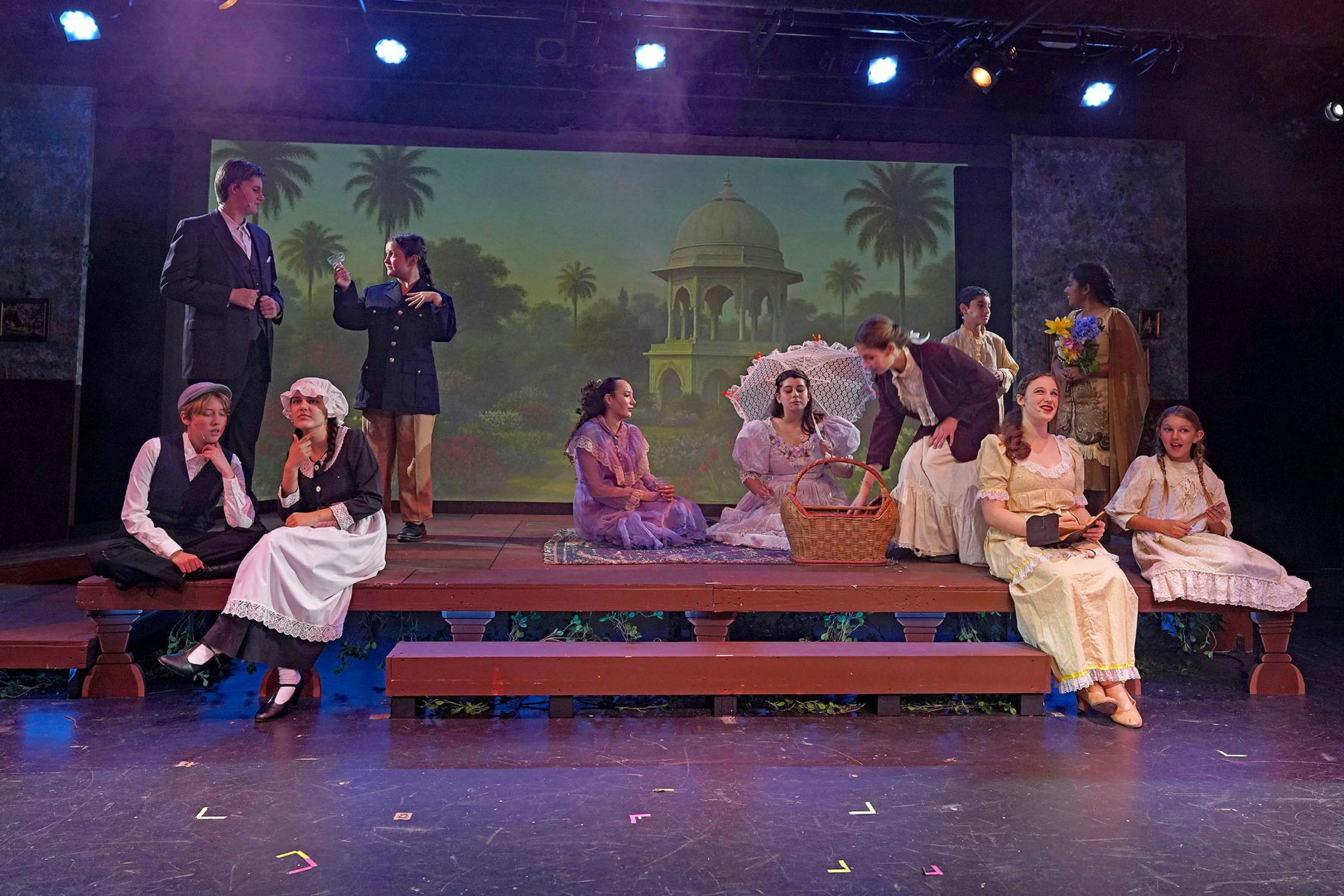 A theater scene with actors in period costumes, sitting and standing on stage, with a scenic backdrop of a garden and pavilion.