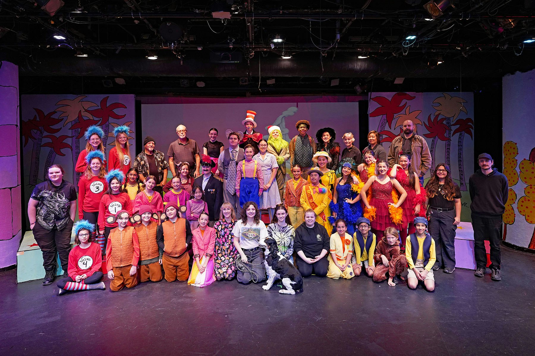 A large group of actors, including children and adults, poses on stage in colorful costumes, likely after a theater performance.